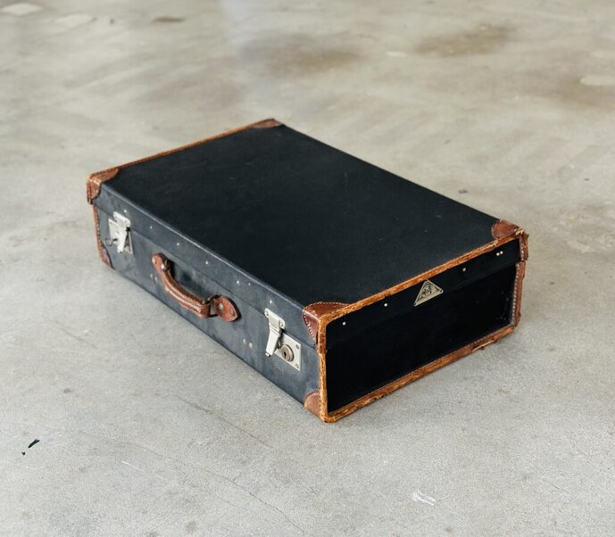 Bildnachweis: Künstlerhaus Stuttgart, 2023. [Bildbeschreibung: Ein schwarzer Koffer mit brauner Zierleiste, braunem Griff und silbernen Verschlussschnallen liegt flach auf einem grauen Betonboden.]